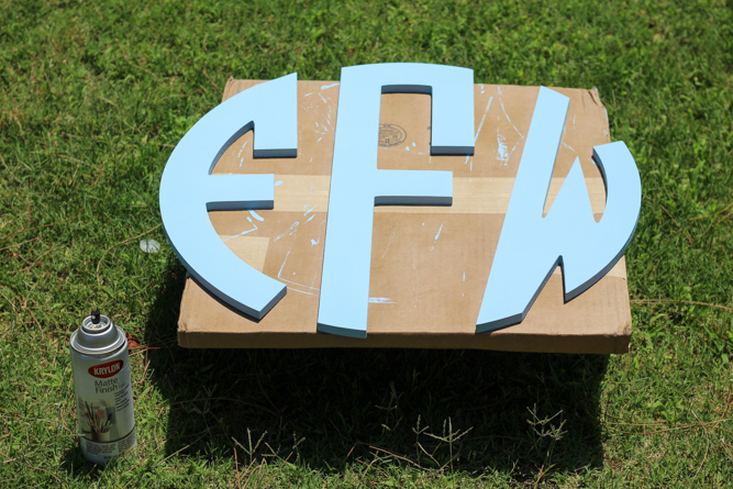 a wood monogram painted light blue that is sitting on cardboard box in the grass while the paint dries