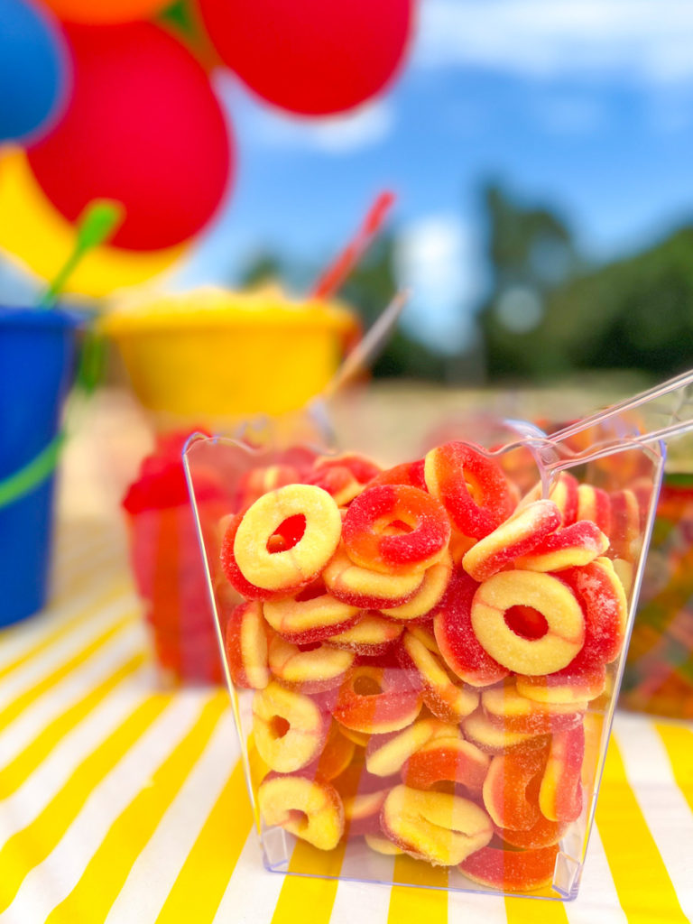 peach ring candy displayed in a clear container on top of a yellow and white stripe tablecloth 