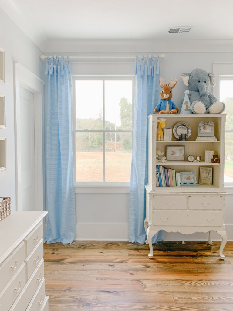blue and white gingham tie-top curtains hanging on a curtain rod in a nursery next to a bookcase decorated with books and stuffed animals 