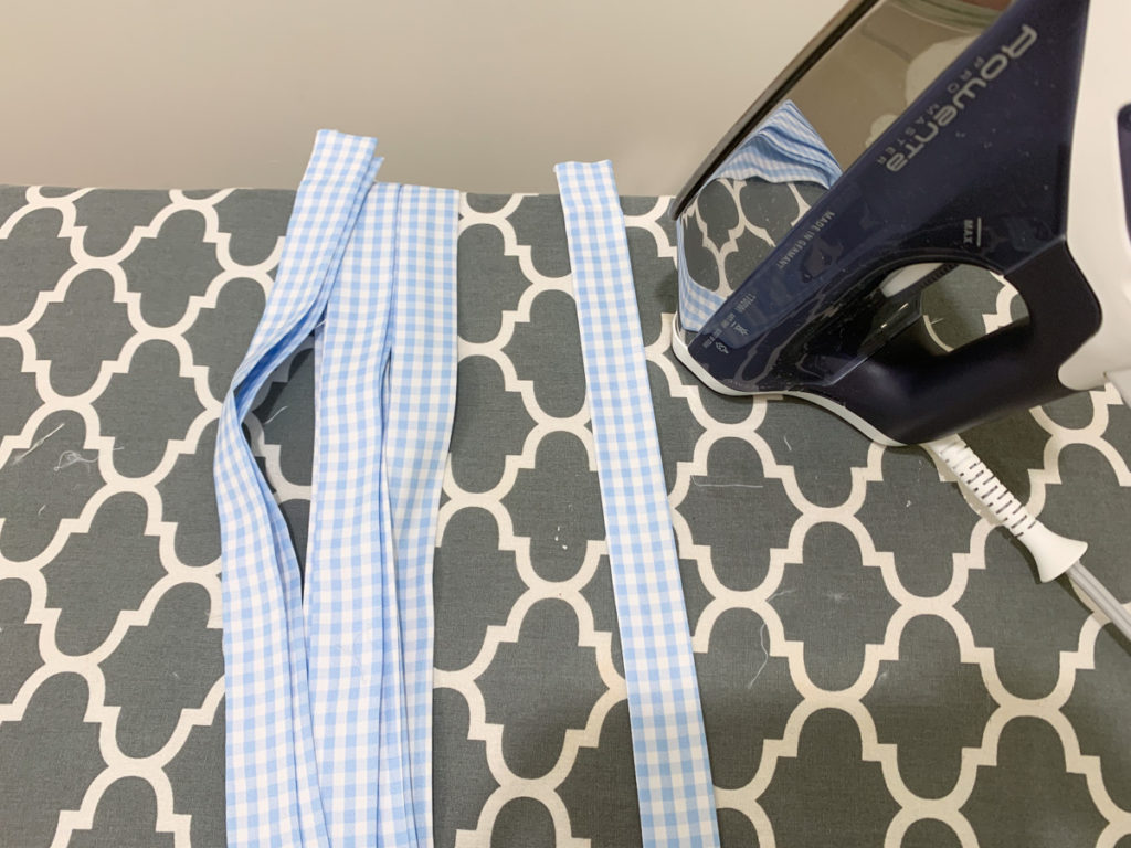 blue and white gingham fabric cut into strips for tie top curtains and sitting on an ironing board next to an iron