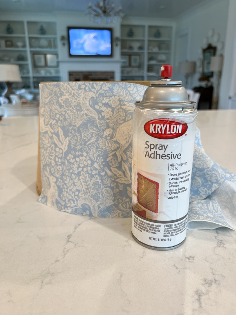 a can of spray adhesive sitting in from of a lampshade that is being covered in fabric for a diy craft project 