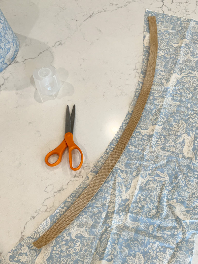 scissors next to a piece of edge banding being wrapped in fabric for a diy project to cover a lampshade with fabric 