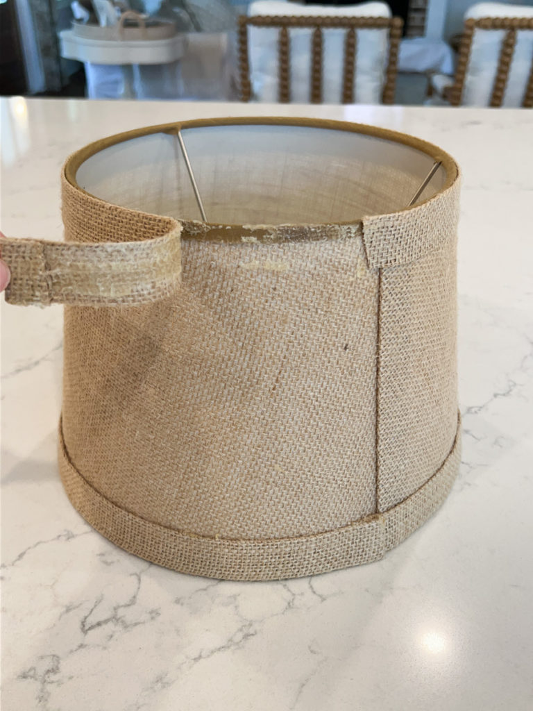 the top edge banding being pulled away and removed from a burlap lampshade for a DIY project to cover a lampshade with fabric 