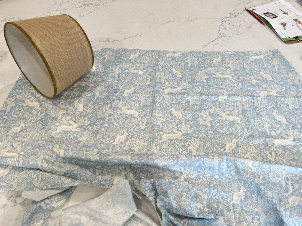 a burlap lampshade sitting on top of a piece of blue and white bunny fabricfor a DIY project to cover a lampshade with fabric 