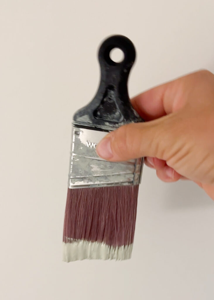 hand holding a paint brush that has been dipped in sage green paint