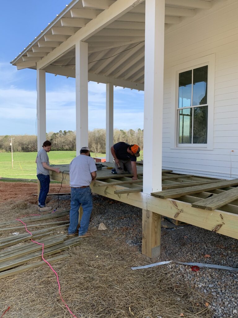 three men installing a wood porch floor on a white farmhouse during construction 